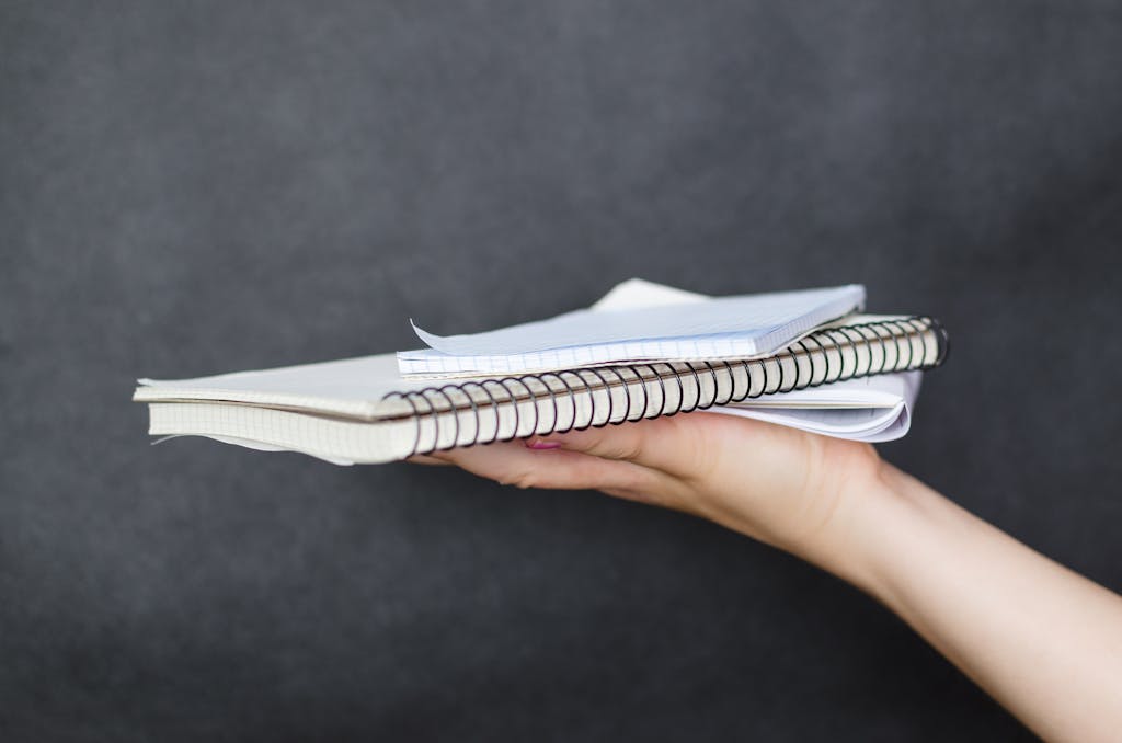 Close-up image of a woman's hand holding a stack of spiral-bound notebooks and papers against a dark background.