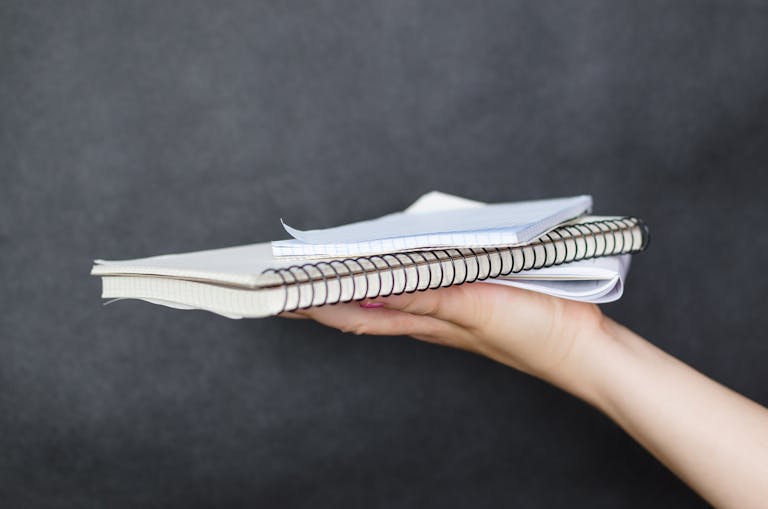 Close-up image of a woman's hand holding a stack of spiral-bound notebooks and papers against a dark background.