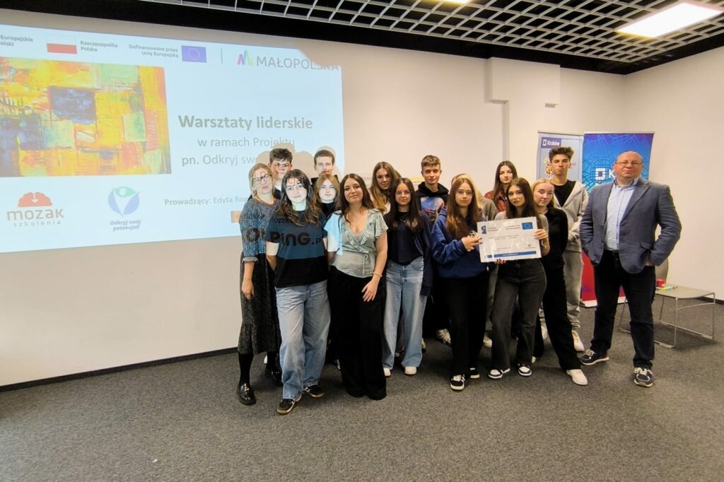Grupa młodych osób pozuje wspólnie do zdjęcia w sali szkoleniowej na tle ekranu z napisem „Warsztaty liderskie w ramach projektu Odkryj swój potencjał”. Obok grupy stoją prowadząca i mężczyzna w garniturze.