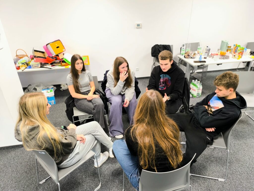 Sześć młodych osób siedzi w kręgu na krzesłach w sali warsztatowej i prowadzi rozmowę. W tle stół z napojami i przekąskami oraz kolorowe materiały plastyczne.