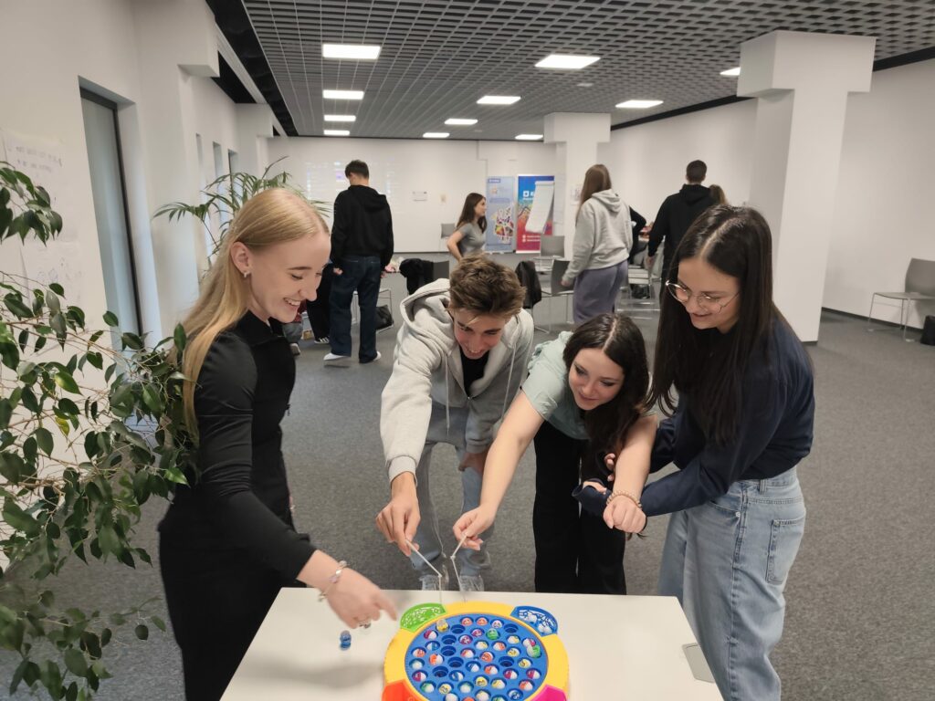 Czworo młodych uczestników warsztatów bawi się przy stole w grę zręcznościową z kolorowymi rybkami. W tle widać innych uczestników zajęć w przestronnej sali szkoleniowej.