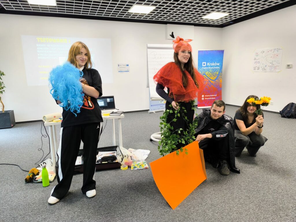 Czworo uczestników warsztatów pozuje w sali szkoleniowej w kolorowych, improwizowanych strojach. Jedna osoba trzyma niebieskie włóczkowe rekwizyty, inna ma czerwoną ozdobę na głowie i sztuczne pnącze, pozostali trzymają kwiaty i pomarańczowy karton.