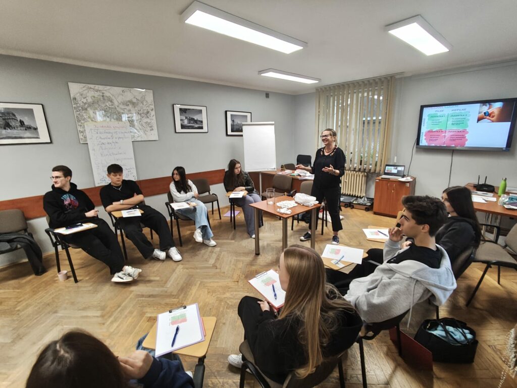 Grupa młodych osób siedzi w kręgu w sali szkoleniowej i słucha kobiety prowadzącej warsztaty. Na ekranie widoczna jest prezentacja, a na tablicy zapisane punkty zajęć.