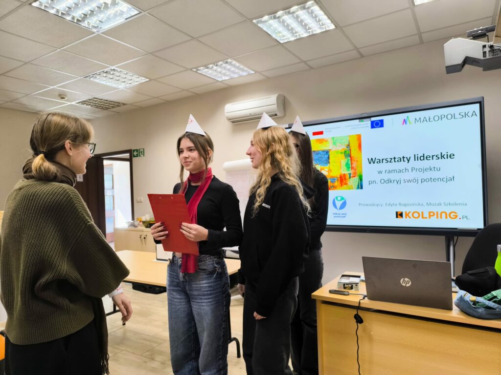 Trzy młode osoby biorą udział w warsztatach liderskich w sali szkoleniowej. Jedna z uczestniczek trzyma czerwoną teczkę i rozmawia z prowadzącą. W tle ekran z napisem „Warsztaty liderskie w ramach projektu Odkryj swój potencjał” oraz logotypami Małopolski, Unii Europejskiej i Kolping.pl.