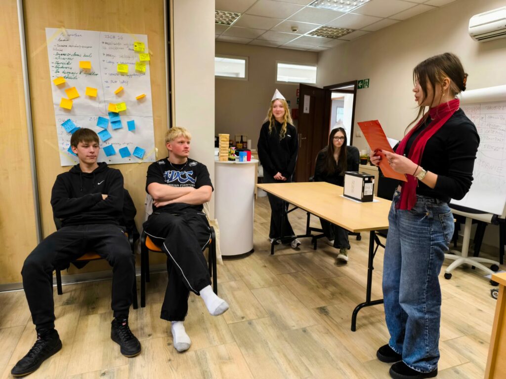 Grupa młodych osób uczestniczy w warsztatach liderskich w sali szkoleniowej. Jedna z uczestniczek stoi i czyta z czerwonej teczki, pozostali siedzą lub stoją, słuchając. W tle tablica z kolorowymi karteczkami i notatkami z zajęć.