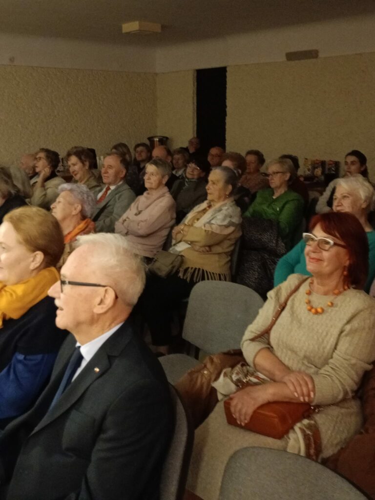 Widownia złożona głównie z osób starszych siedzi na krzesłach i obserwuje występ. Ludzie patrzą przed siebie z zaciekawieniem i uśmiechem. W pomieszczeniu panuje ciepłe oświetlenie.