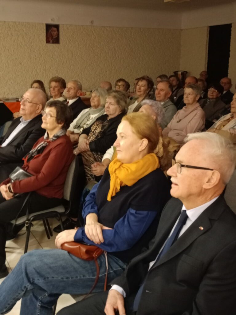 Grupa widzów siedzi na widowni, słuchając programu. Część osób ma założone eleganckie ubrania, szale lub ozdobne korale. Wszyscy patrzą w stronę sceny i wydają się skupieni oraz zaangażowani.