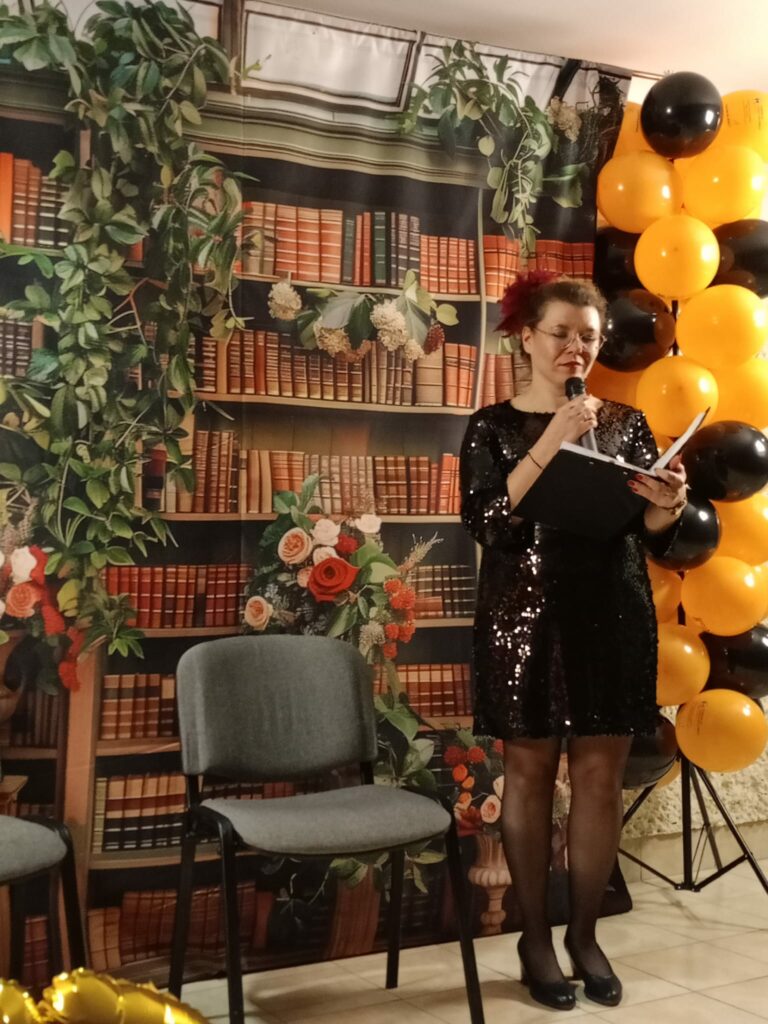 Kobieta w czarnej, błyszczącej sukience stoi na scenie i mówi do mikrofonu, trzymając otwarty segregator. W tle dekoracja przedstawiająca regały z książkami i kompozycje kwiatowe oraz kolumny balonów w kolorach pomarańczowym i czarnym.