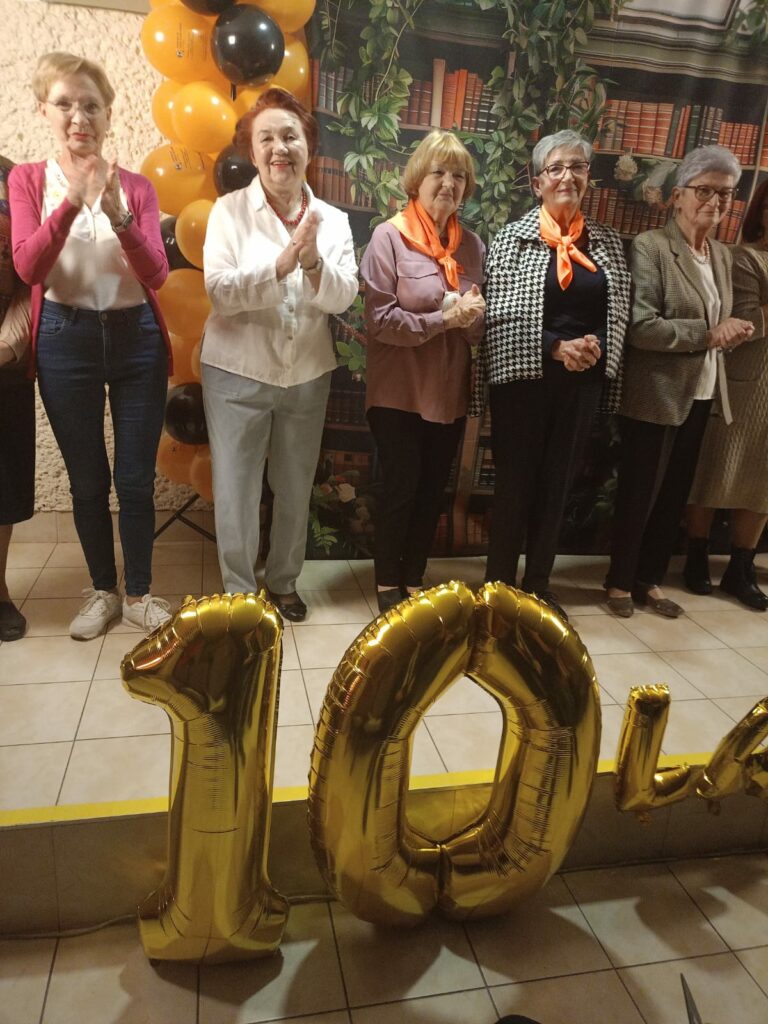 Sześć kobiet stoi w rzędzie i bije brawo. Przed nimi znajdują się złote balony w kształcie liczby „10”. W tle dekoracja z bibliotecznymi półkami i balony w kolorach czarnym oraz pomarańczowym.