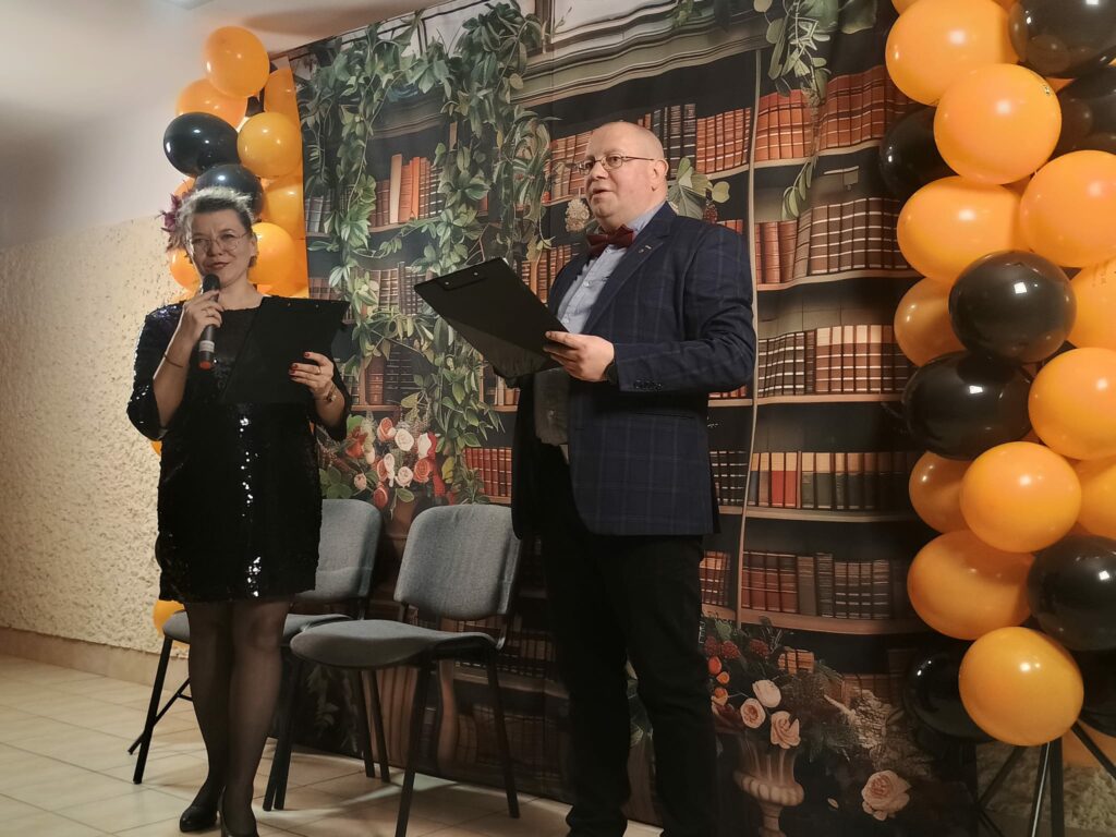 Kobieta i mężczyzna prowadzą część uroczystości, trzymając mikrofony i czarne teczki. Kobieta ma czarną błyszczącą sukienkę, mężczyzna ma granatową marynarkę. Za nimi dekoracja w formie biblioteki oraz balony.