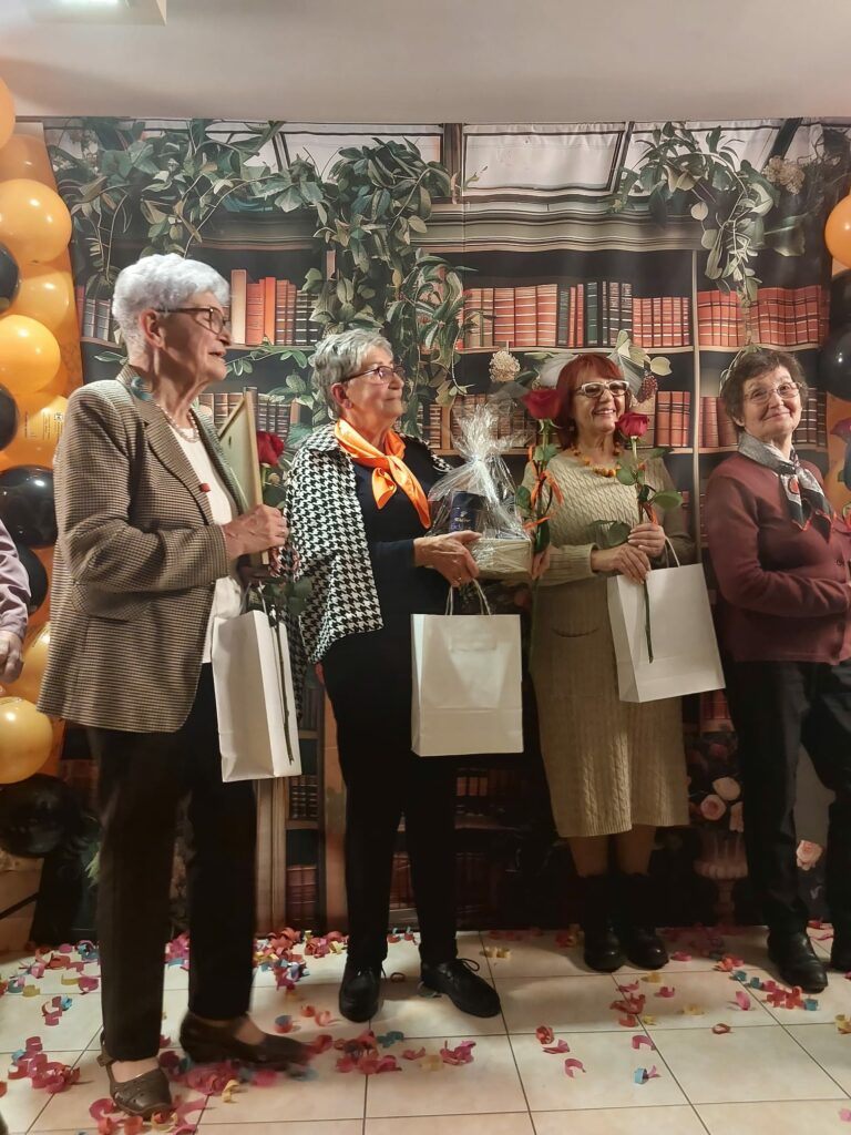 Pięć kobiet stoi w rzędzie, każda trzyma różę oraz biały upominkowy worek. Obok nich stoją balony w czarnym i pomarańczowym kolorze. Na podłodze leżą kolorowe konfetti.