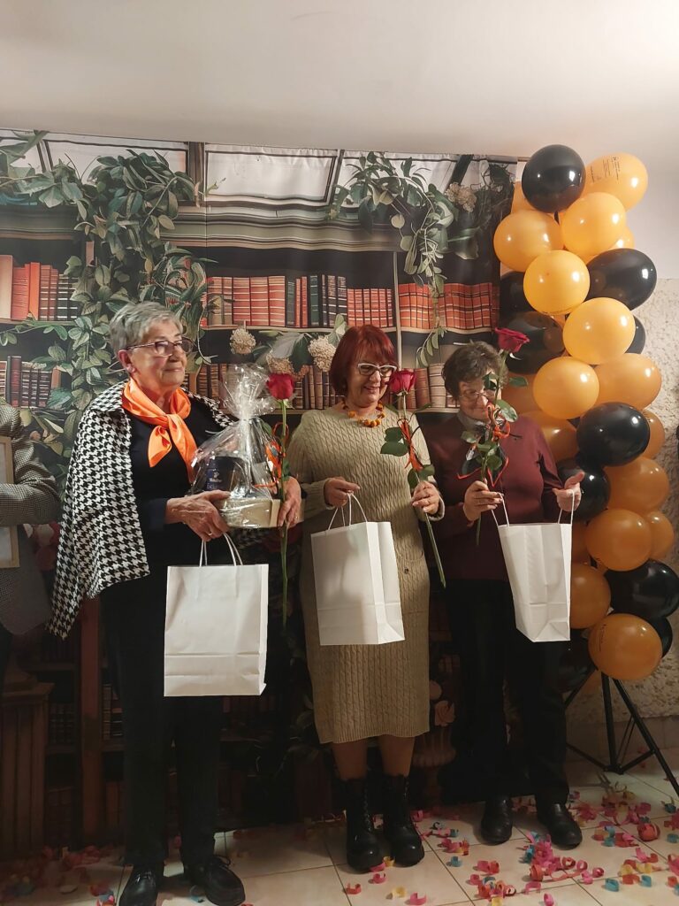Trzy kobiety stoją obok siebie, trzymając róże i białe torby z upominkami. Jedna z kobiet trzyma także przezroczysty prezent ozdobiony celofanem. Tło stanowi dekoracja z motywem biblioteki i balony w czarnym i pomarańczowym kolorze.