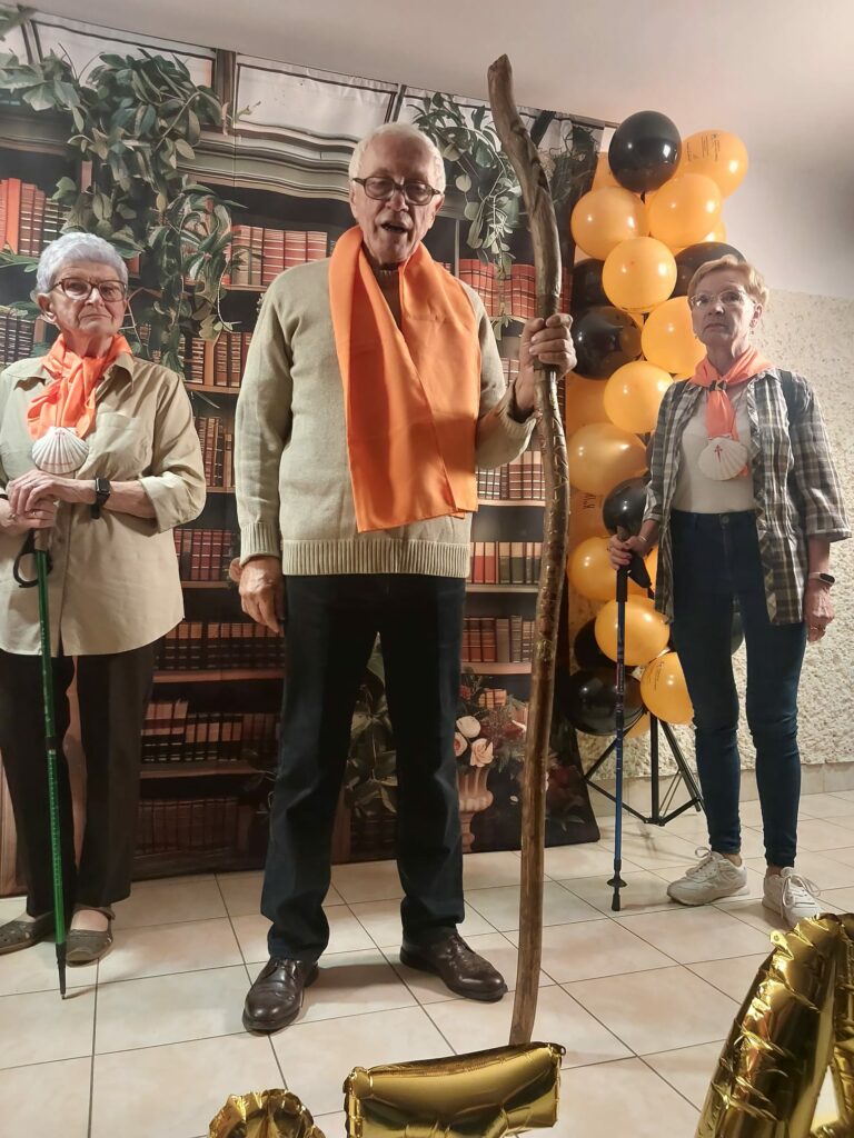 Trzy starsze osoby stoją na scenie. Mężczyzna w środku trzyma długi drewniany kij. Wszyscy mają pomarańczowe chusty na szyjach. W tle znajduje się dekoracja przedstawiająca regał z książkami i balony w czarnym oraz pomarańczowym kolorze.