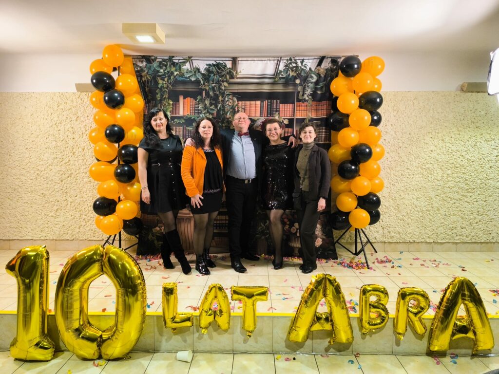 Pięć osób pozuje razem do zdjęcia na tle dekoracji z książkami. Po bokach stoją kolumny z balonów w kolorach czarnym i pomarańczowym. Na podłodze ułożone są złote balony tworzące napis „10 lat ABRA”.