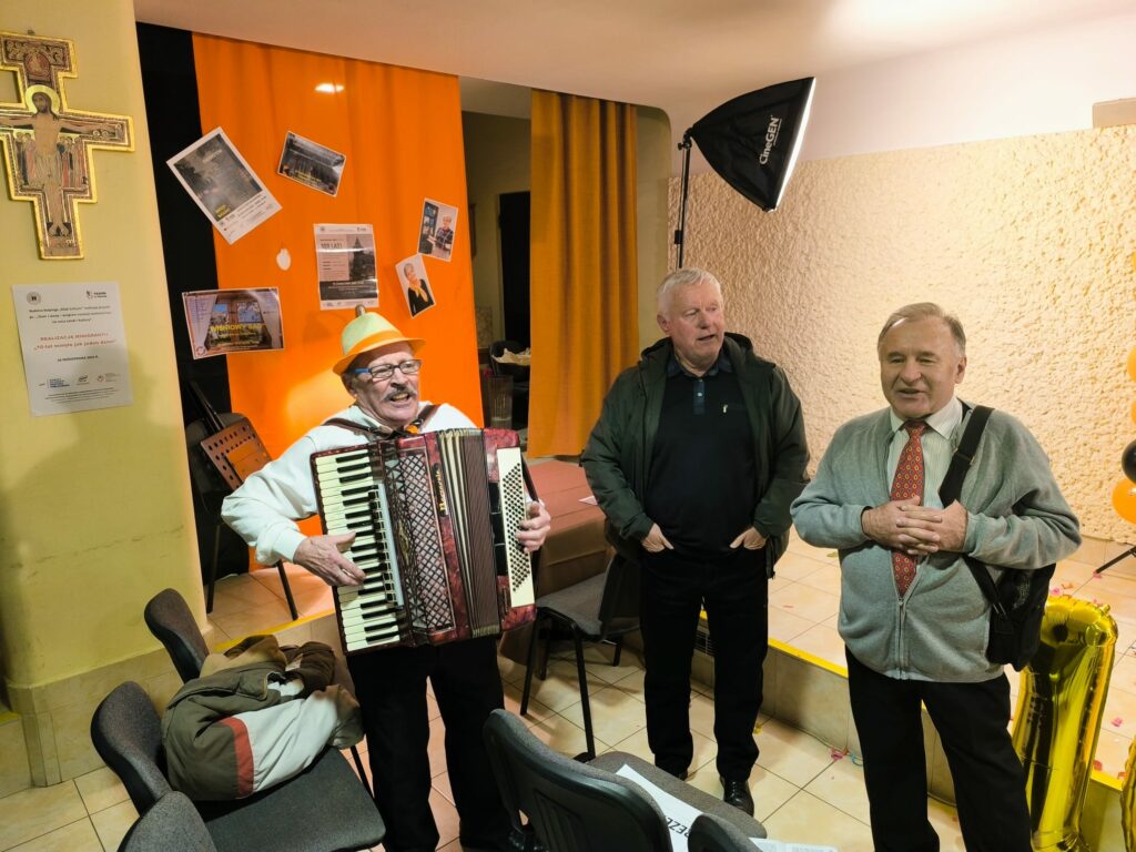 Trzech starszych mężczyzn stoi w pomieszczeniu. Jeden z nich gra na akordeonie i ma na głowie żółty kapelusz. Pozostali dwaj słuchają z uśmiechem. W tle wiszą pomarańczowe zasłony, plakaty oraz lampa do oświetlenia zdjęć.