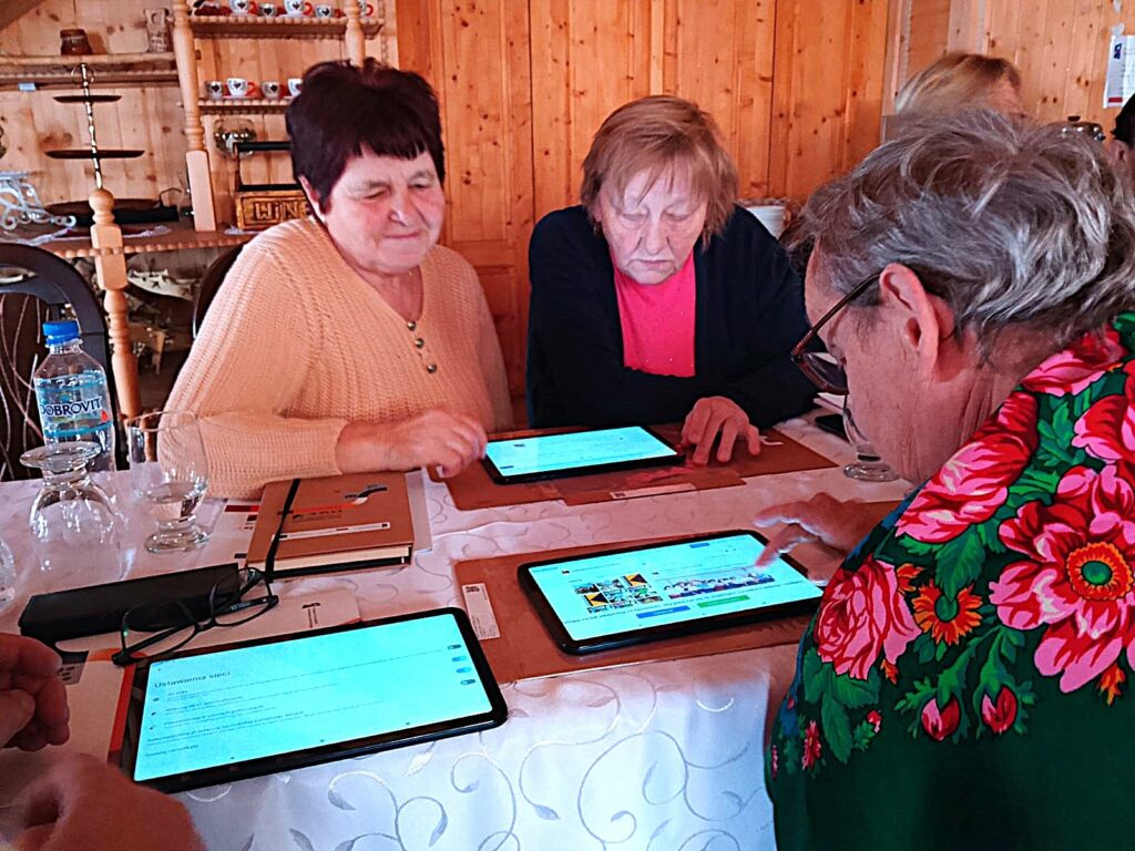 Kilka seniorek siedzi przy stole i wspólnie korzysta z tabletów podczas warsztatów cyfrowych. Uczestniczki skupione są na ekranach urządzeń, ucząc się obsługi aplikacji w przyjaznej, kameralnej przestrzeni.