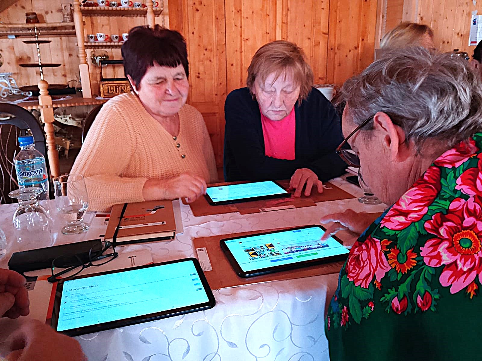 Kilka seniorek siedzi przy stole i wspólnie korzysta z tabletów podczas warsztatów cyfrowych. Uczestniczki skupione są na ekranach urządzeń, ucząc się obsługi aplikacji w przyjaznej, kameralnej przestrzeni.