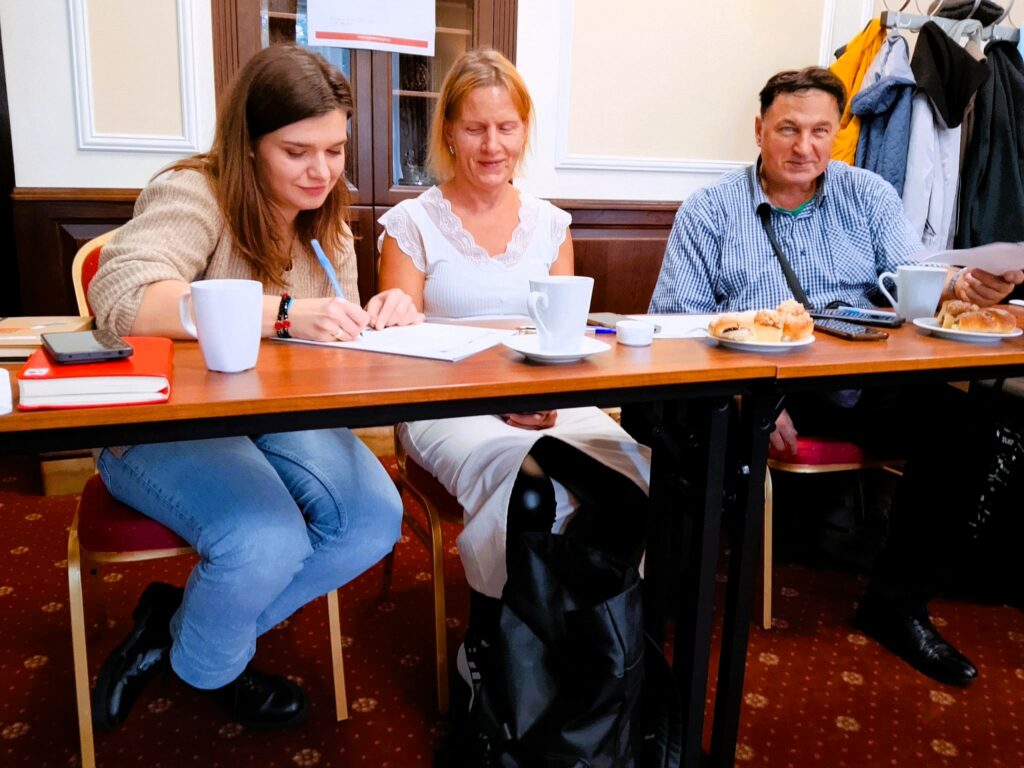 Trzy osoby siedzą przy stole podczas spotkania – młoda kobieta zapisuje notatki, obok niej dwie osoby przeglądają dokumenty. Na stole stoją kubki z kawą i talerzyki z ciastem.