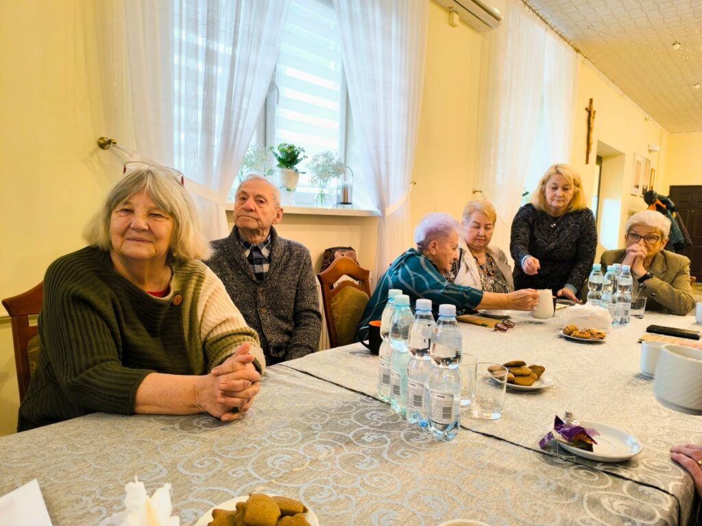 Grupa seniorów i wolontariuszy siedzących przy wspólnym stole w jasnej sali, podczas świątecznego spotkania Rodziny Kolpinga. Na stole woda, ciastka i kubki, uczestnicy rozmawiają w spokojnej, serdecznej atmosferze.