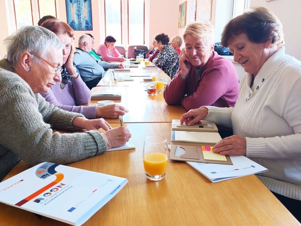 Grupa seniorów siedzi przy długim stole podczas warsztatów lub spotkania. Uczestnicy robią notatki i rozmawiają, na stole leżą materiały szkoleniowe oraz szklanki z sokiem.