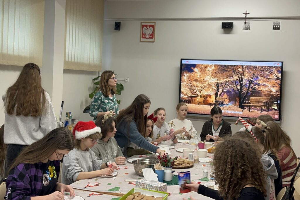 Grupa młodzieży i opiekunów wspólnie dekoruje elementy z piernika przy długim stole, przygotowując świąteczne domki z piernika w sali szkolnej.