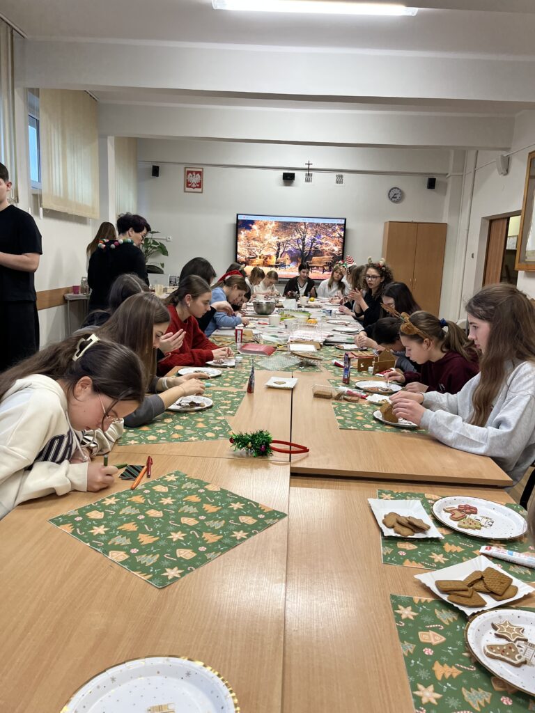 Grupa młodzieży i opiekunów wspólnie dekoruje elementy z piernika przy długim stole, przygotowując świąteczne domki z piernika w sali szkolnej.