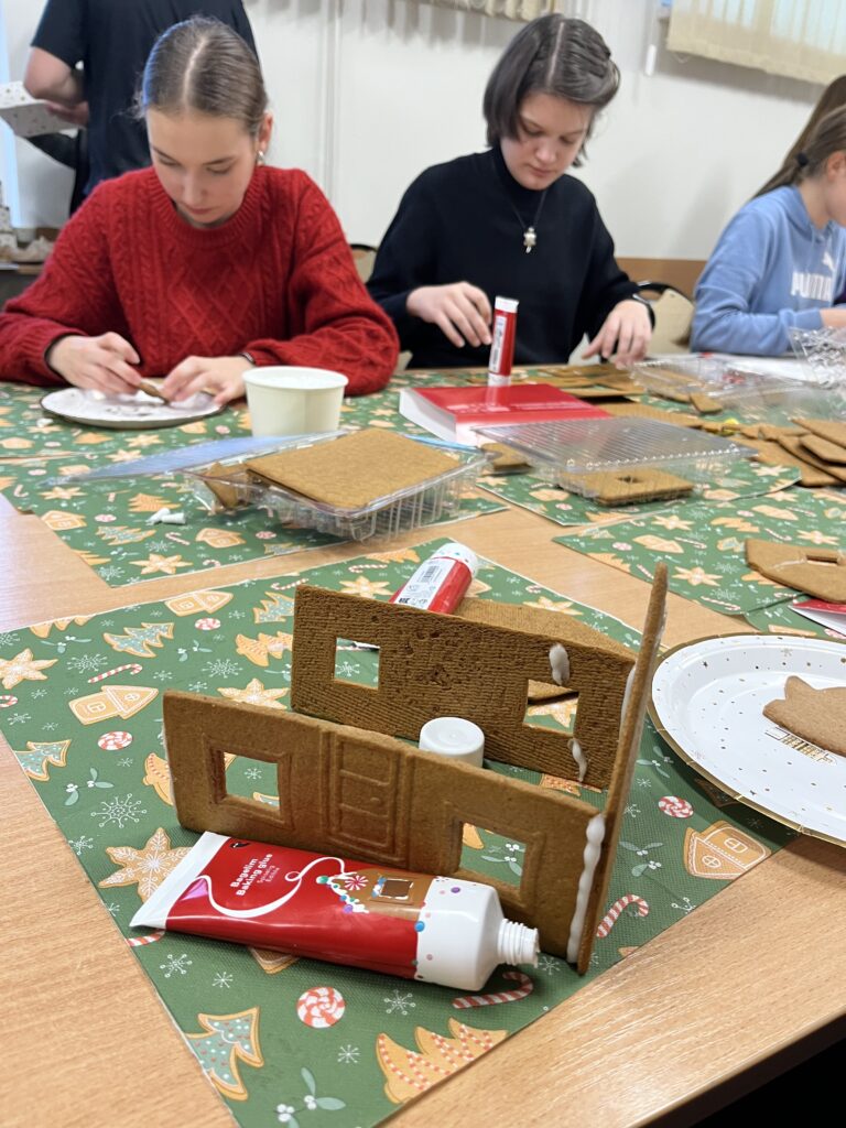 Zbliżenie na składane elementy piernikowego domku na stole, obok tubki z lukrem i talerzyków, w tle młodzież pracująca nad dekoracjami.