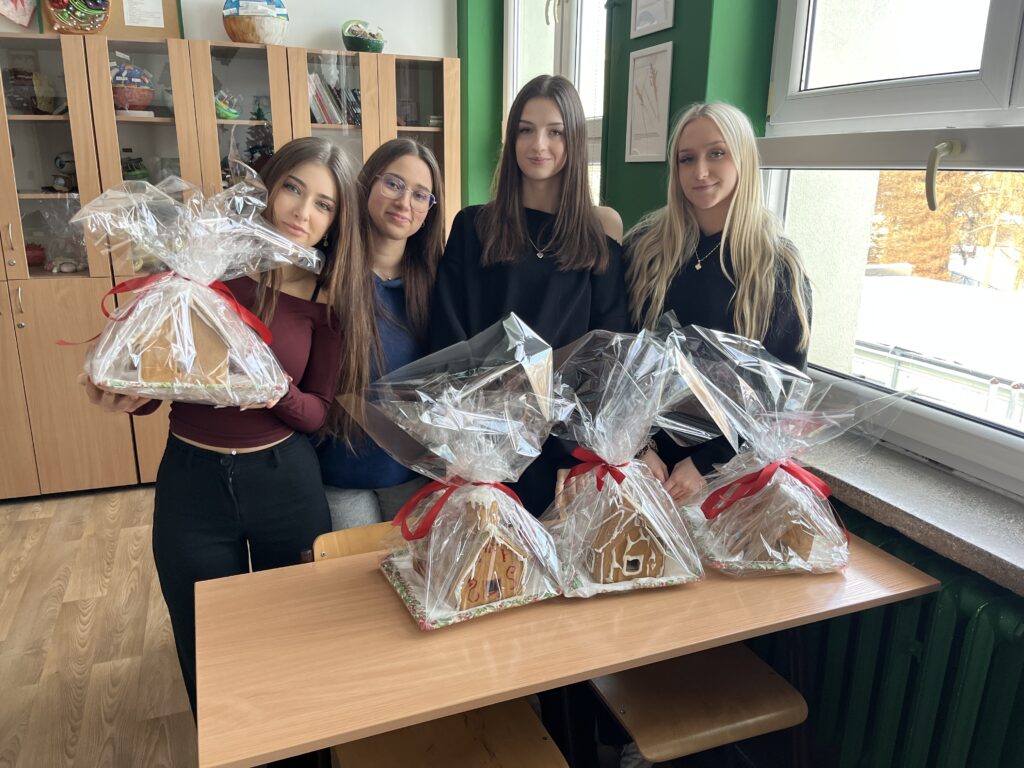 Cztery młode osoby stoją przy szkolnej ławce i prezentują zapakowane w folię celofanową domki z piernika przewiązane czerwoną wstążką.
