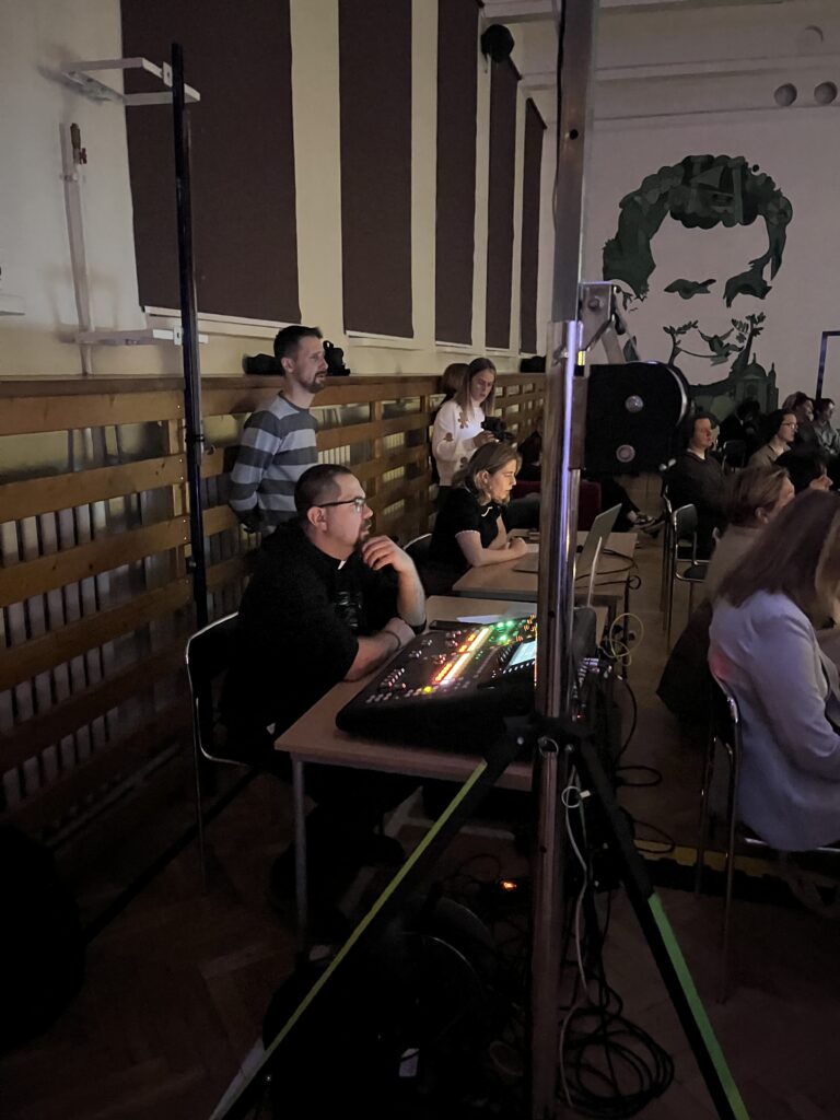 tanowisko techniczne z mikserem dźwięku podczas wydarzenia w sali gimnastycznej, obok siedzą osoby obsługujące nagłośnienie i oświetlenie.