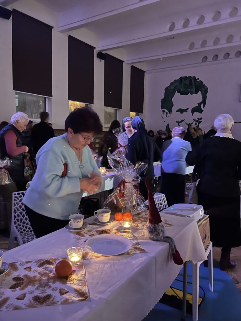 Starsze osoby i uczestnicy spotkania rozmawiają i częstują się przy stołach podczas świątecznego wydarzenia w sali, w tle dekoracje i goście.