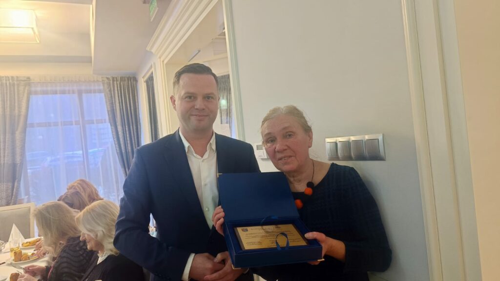 „Dwie osoby stojące w jasnym wnętrzu podczas uroczystego spotkania. Kobieta trzyma otwarte etui z pamiątkową tabliczką lub dyplomem, obok niej mężczyzna w garniturze. W tle widoczni uczestnicy siedzący przy stole.”