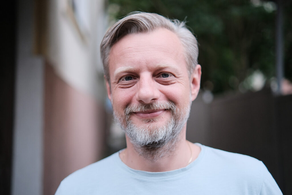 Portret uśmiechniętego mężczyzny z jasnymi włosami i krótką siwą brodą, patrzącego w obiektyw. Stoi na zewnątrz, a w tle widać rozmytą zieleń i fragment budynku.