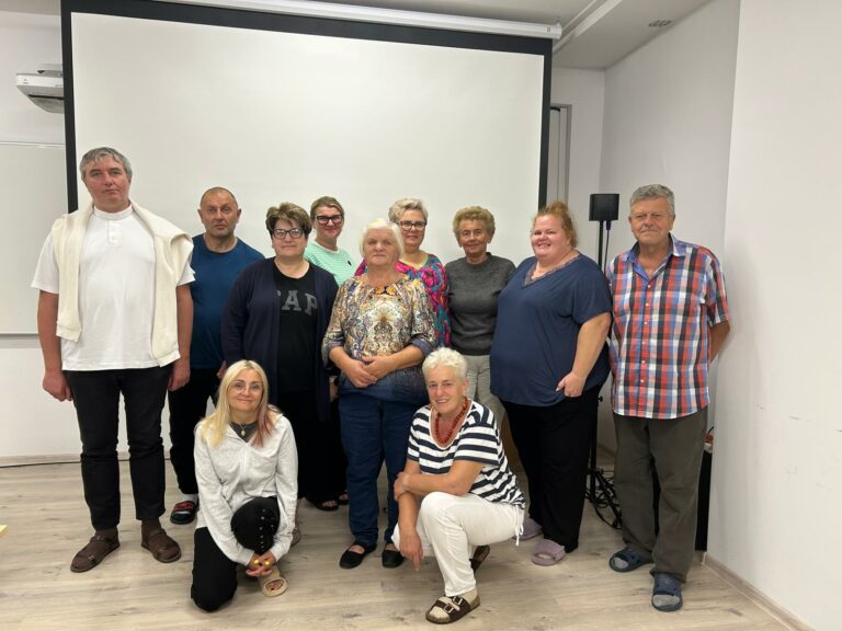 Grupa jedenastu dorosłych osób stoi i klęczy do wspólnego zdjęcia w jasnej sali szkoleniowej. W tle widoczny jest ekran projekcyjny. Uczestnicy są ubrani na co dzień i patrzą w stronę aparatu.