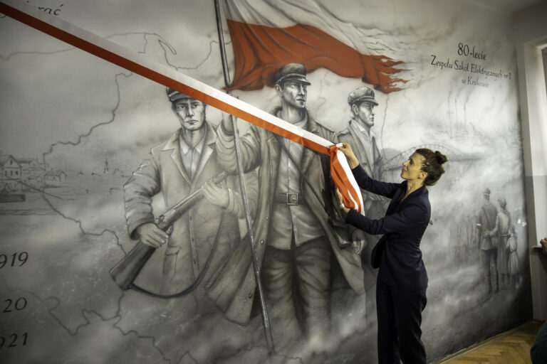 „Kobieta w eleganckim stroju odsłania czerwono-białą wstęgę na ścianie z muralem przedstawiającym trzech uzbrojonych powstańców oraz dużą flagę w barwach Polski. Na muralu widoczny jest także napis o 80-leciu Zespołu Szkół Elektrycznych nr 1 w Krakowie oraz zarys mapy i elementy miejskiego krajobrazu w tle.”