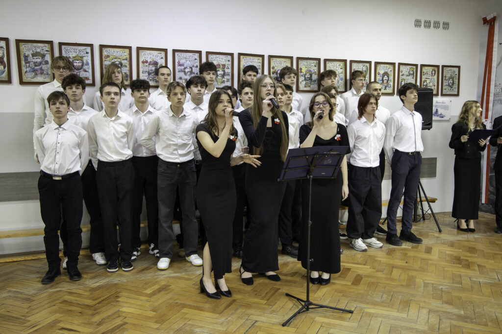 „Grupa uczniów stoi w rzędach, tworząc chór podczas występu. Na pierwszym planie trzy dziewczyny w czarnych strojach śpiewają do mikrofonów przy pulpicie na nuty. W tle widoczni są pozostali uczniowie w białych koszulach oraz ściana z oprawionymi ilustracjami.”