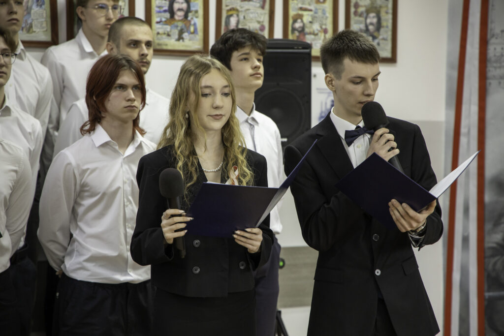 „Dwoje uczniów prowadzi uroczystość, stojąc z mikrofonami i czytając z kartek. Chłopak w garniturze i muszce przemawia do mikrofonu, a dziewczyna obok trzyma mikrofon i notatki. W tle stoi grupa uczniów w białych koszulach oraz ściana z oprawionymi ilustracjami.”