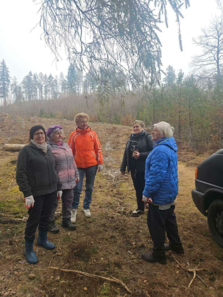 Pięć starszych kobiet stoi na leśnej polanie i rozmawia. Ubrane są w ciepłe kurtki, czapki i rękawiczki robocze, co sugeruje udział w pracach terenowych. W tle widać młode drzewa i fragment lasu, a z prawej strony zaparkowany samochód.