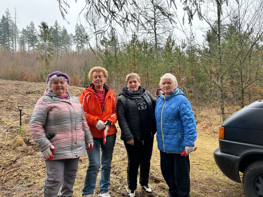 Cztery starsze kobiety stoją razem na skraju lasu, ubrane w ciepłe kurtki i rękawiczki robocze. Jedna z nich trzyma narzędzie ogrodowe, a wszystkie wyglądają na zaangażowane w prace terenowe, prawdopodobnie sprzątanie lub porządkowanie terenu. W tle widać młode drzewa i leśny krajobraz wczesnej wiosny lub późnej jesieni, a z prawej strony fragment zaparkowanego samochodu.
