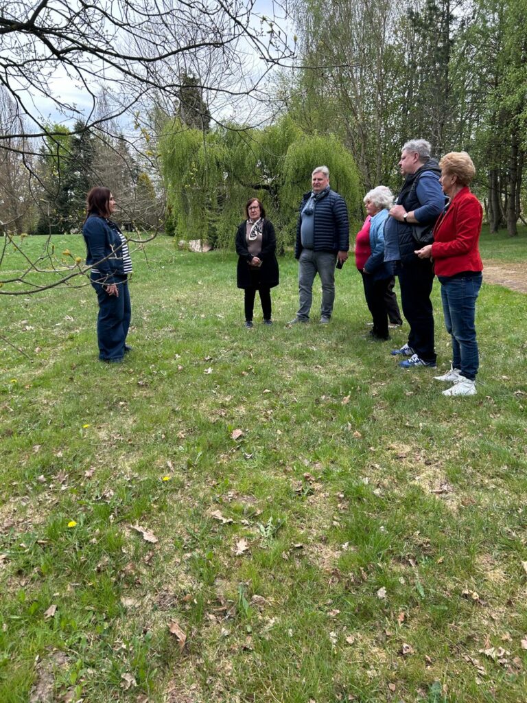 Grupa kilku osób stoi na polanie w parku lub ogrodzie i słucha kobiety prowadzącej spotkanie. Uczestnicy są zwróceni w jej stronę, a wokół widać zieloną trawę, drzewa oraz wiosenną przyrodę. Zdjęcie przedstawia spokojny moment wspólnego spaceru lub warsztatów na świeżym powietrzu.