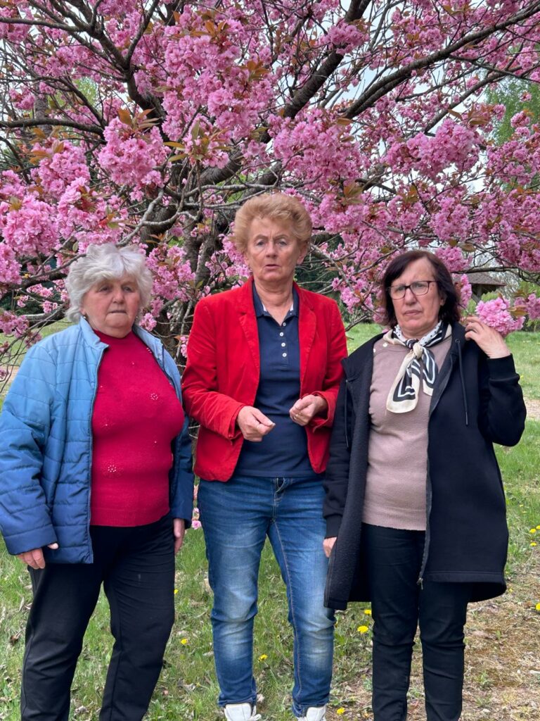 Trzy starsze kobiety stoją pod drzewem obsypanym intensywnie różowymi kwiatami. W tle widać wiosenną zieleń, a kobiety patrzą w stronę aparatu. Jedna ma czerwoną marynarkę, druga niebieską kurtkę, a trzecia czarną bluzę i apaszkę. Atmosfera zdjęcia jest spokojna i wiosenna.