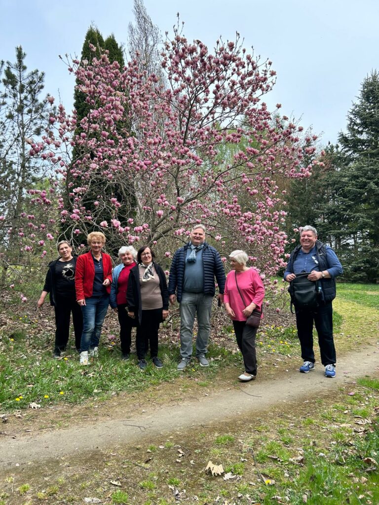 Grupa siedmiu osób stoi na ścieżce i trawie przed dużym drzewem magnolii obsypanym różowymi kwiatami. Uczestnicy uśmiechają się i pozują do zdjęcia w otoczeniu wiosennej zieleni oraz innych drzew. Atmosfera zdjęcia jest radosna i spokojna.