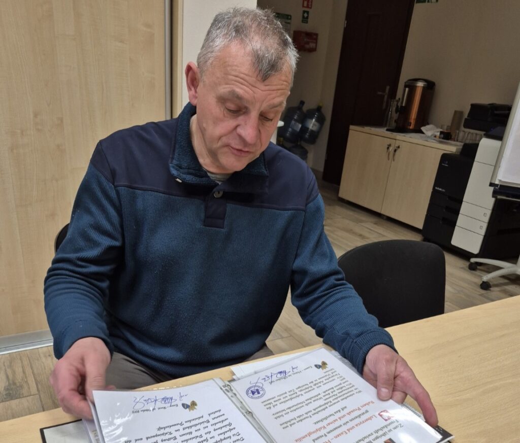 Mężczyzna siedzi przy stole w biurze i przegląda otwarty segregator z dokumentami oraz dyplomami związanymi z działalnością organizacji. W tle widoczne są szafki, ekspres do kawy i sprzęt biurowy. Skupiony na lekturze dokumentów, trzyma kartki obiema rękami.