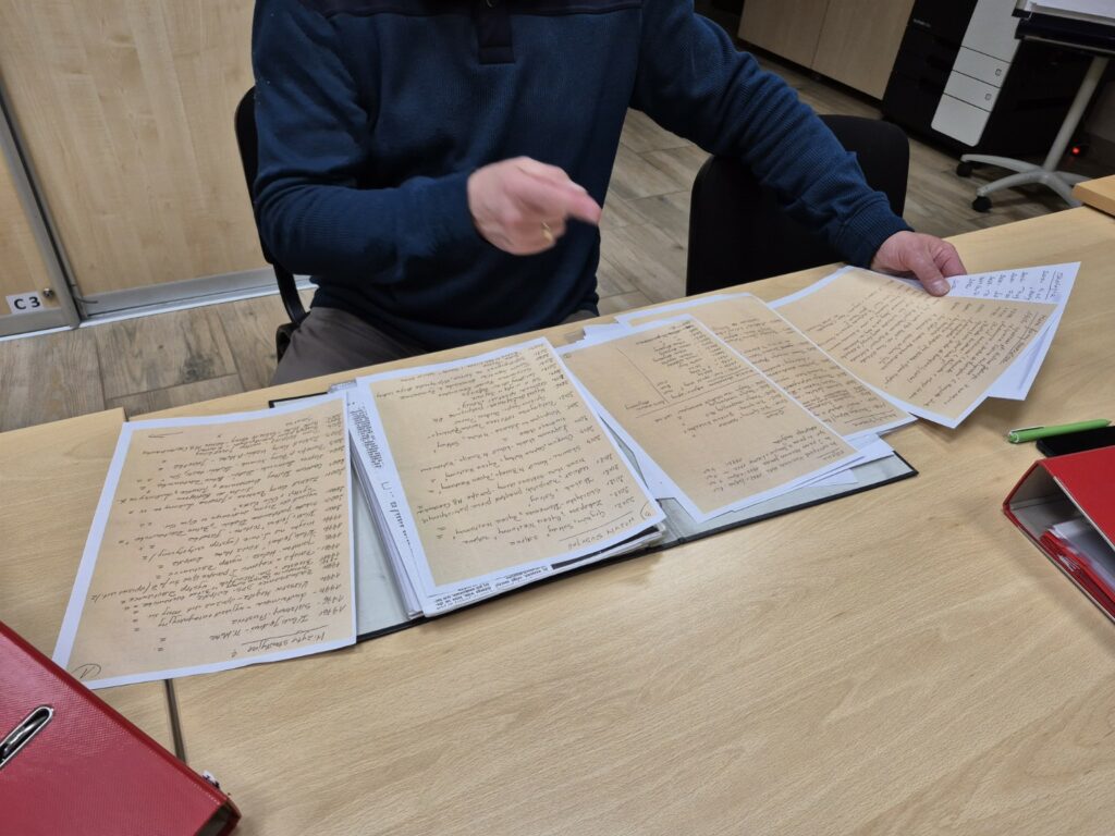 Mężczyzna siedzi przy stole i przegląda archiwalne dokumenty umieszczone w segregatorze. Na blacie rozłożone są kopie odręcznie zapisanych notatek i zestawień, które wskazuje dłonią podczas omawiania ich zawartości. Zdjęcie przedstawia pracę z materiałami dokumentującymi historię i działalność organizacji.