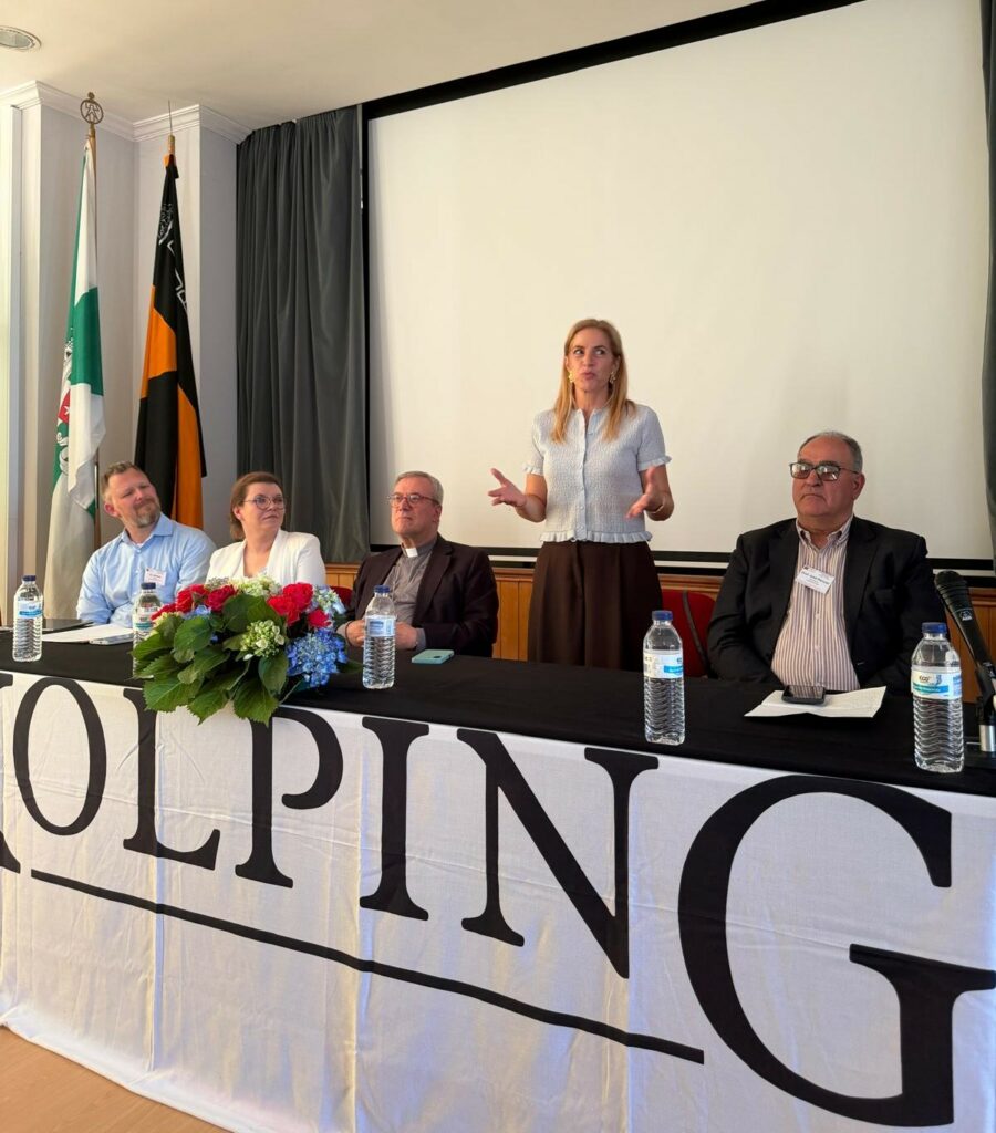 Kobieta przemawia do uczestników Zgromadzenia Kontynentalnego Kolping Europa. Za stołem prezydialnym siedzą przedstawiciele organizacji, a przed nimi znajdują się mikrofony, woda i kompozycja kwiatowa.