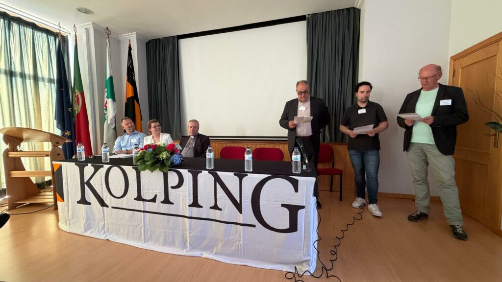 Sala konferencyjna podczas Zgromadzenia Kontynentalnego Kolping Europa. Przy stole prezydialnym siedzą członkowie władz organizacji, a trzech uczestników stojących obok odczytuje przygotowane wystąpienia. Na stole znajduje się baner z napisem „KOLPING”.