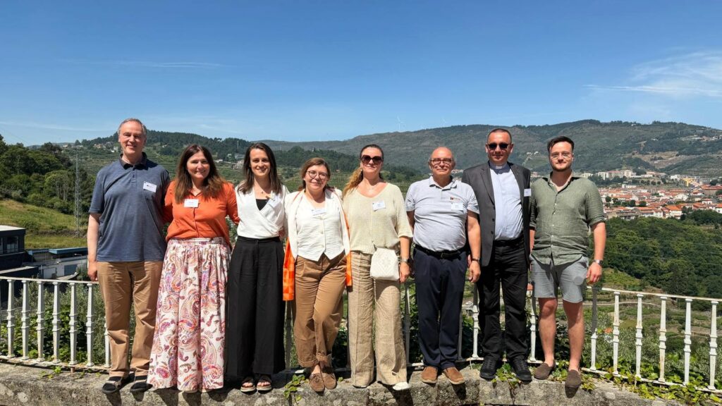 Osiem osób stoi obok siebie na tarasie z panoramą gór i miasta w tle. To przedstawiciele Zarządu KOLPING Europa biorący udział w Zgromadzeniu Kontynentalnym Kolping Europa w Portugalii.