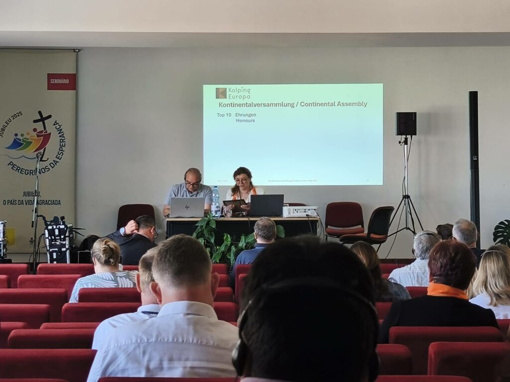 Uczestnicy Zgromadzenia Kontynentalnego Kolping Europa siedzą na sali konferencyjnej i słuchają prezentacji. Na ekranie wyświetlony jest slajd dotyczący działalności Kolping Europa, a prowadzący pracują przy komputerach.
