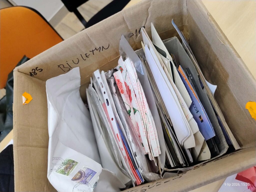 Kartonowe pudełko wypełnione listami, kopertami, albumami i dokumentami archiwalnymi. Na brzegu pudełka widoczny napis „Biuletyny”. Materiały są posegregowane i przygotowane do przeglądu oraz opracowania.