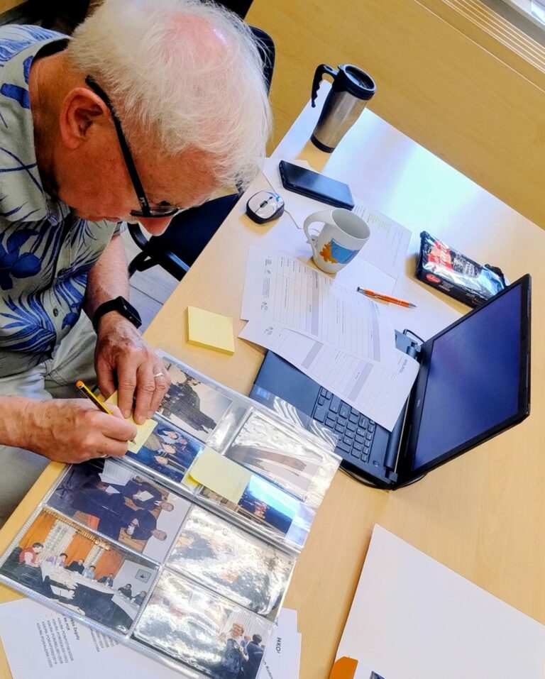 Mężczyzna przegląda album ze zdjęciami podczas pracy nad archiwum. Na stole leżą laptopy, dokumenty, raport działalności KOLPING Polska oraz fotografie, które są identyfikowane i opisywane.