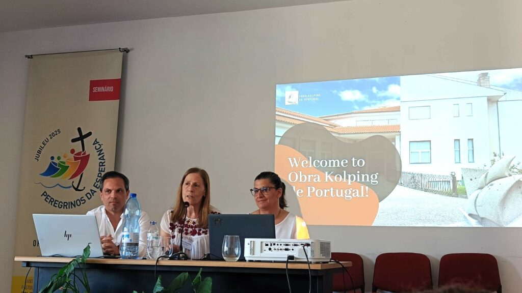 Trzy osoby siedzą przy stole konferencyjnym i prezentują działalność Obra Kolping de Portugal. Za nimi wyświetlony jest slajd z napisem „Welcome to Obra Kolping de Portugal!”, a obok znajduje się baner Jubileuszu 2025.